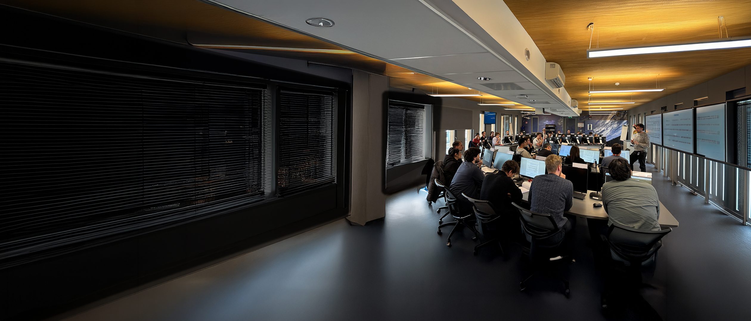 Training room in session — seated group at workstations, lit ceiling, dark interior. Mirrored version with the third-party signage removed. Keep the seated group and the lit ceiling in the crop; a centre crop lands on empty floor.